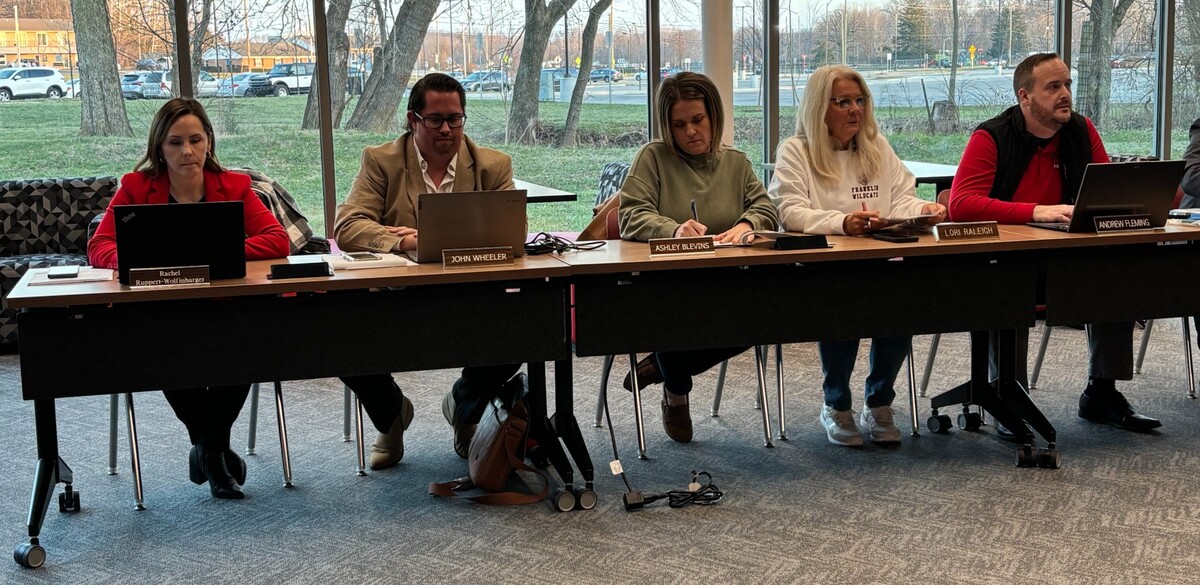 A group of five people sitting at tables in front of windows facing the camera.